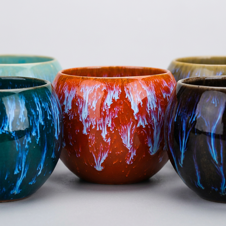 A group of ceramic cups with marbled, swirled glazes in various colors, including teal, orange with white, dark blue, and black with blue, arranged on a light gray background.