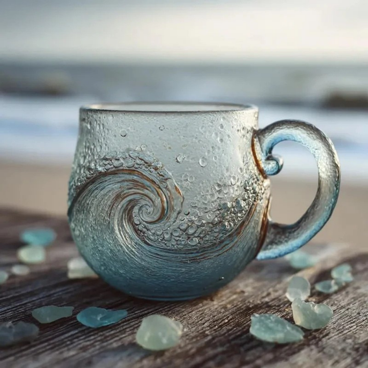 A frosted glass mug with a blue-gray wave design and condensation droplets, set on a wooden surface with scattered sea glass stones, with a blurred beach and ocean background.