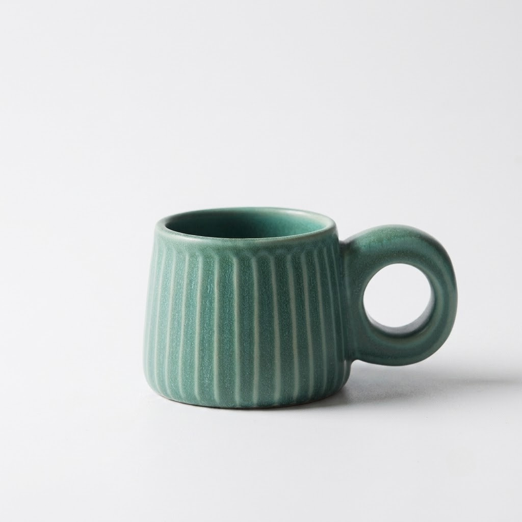A small ceramic mug with a rounded shape, vertical ribbed texture, and a loop handle, finished in a matte sage green glaze, set against a plain light gray background.