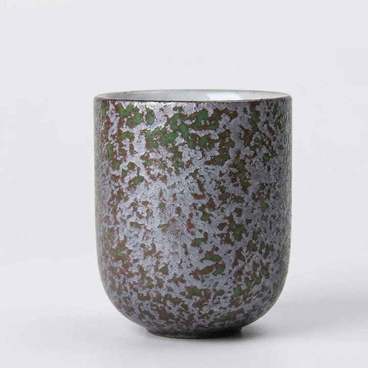 A ceramic cup with a textured, speckled glaze in metallic silver, olive green, and dark brown, set against a plain light gray background.