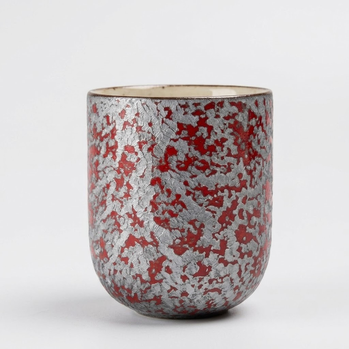 A small ceramic cup with a textured, crackled metallic silver exterior featuring red speckles, and a cream-colored interior, shown on a light gray background.