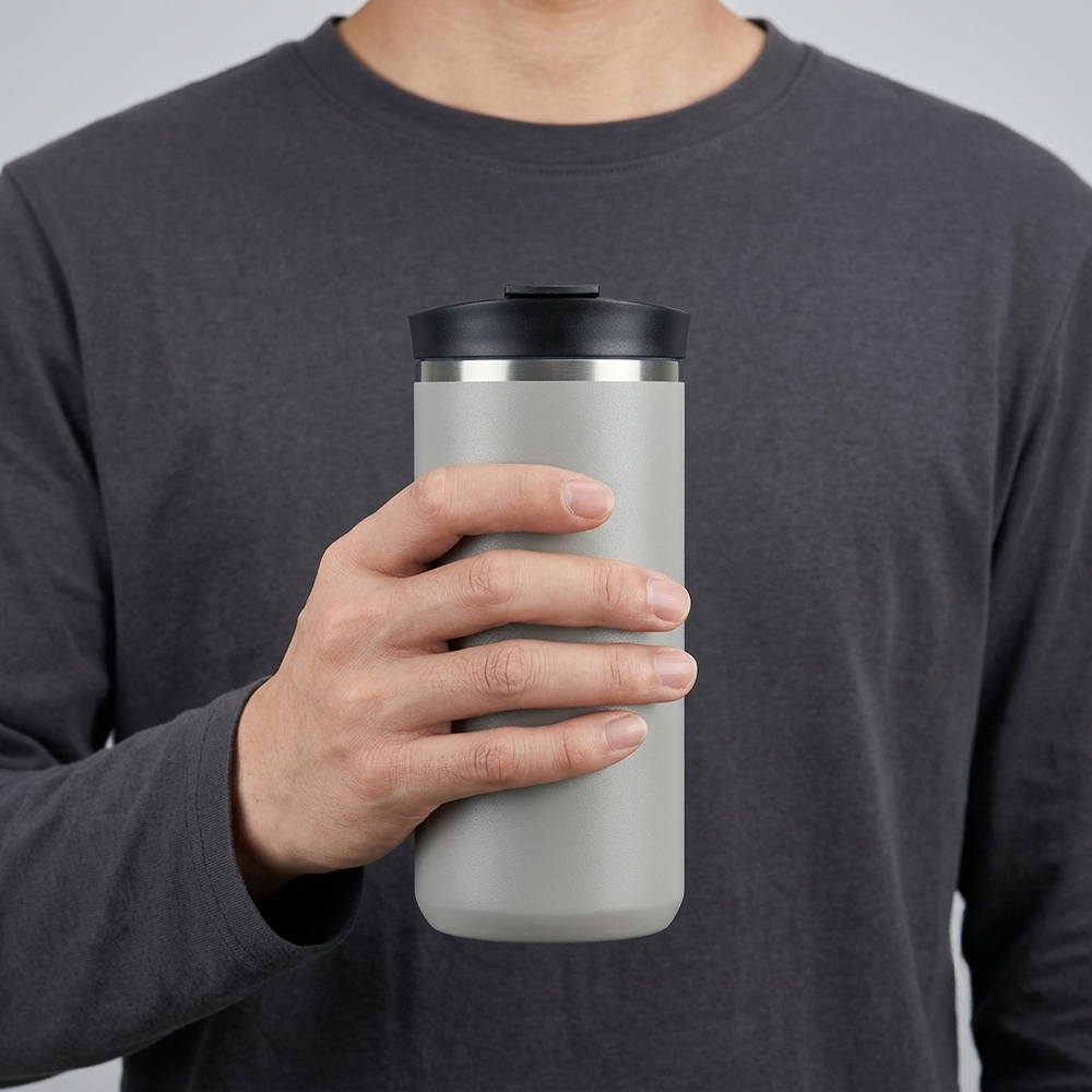 A person in a dark gray long-sleeve shirt holds a matte light gray insulated travel tumbler with a black lid and a thin silver metallic band near the top, against a plain light gray background.