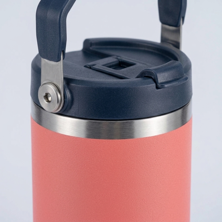 A close-up of a coral pink insulated water bottle with a dark navy blue lid and carry handle, featuring a stainless steel band around the neck.