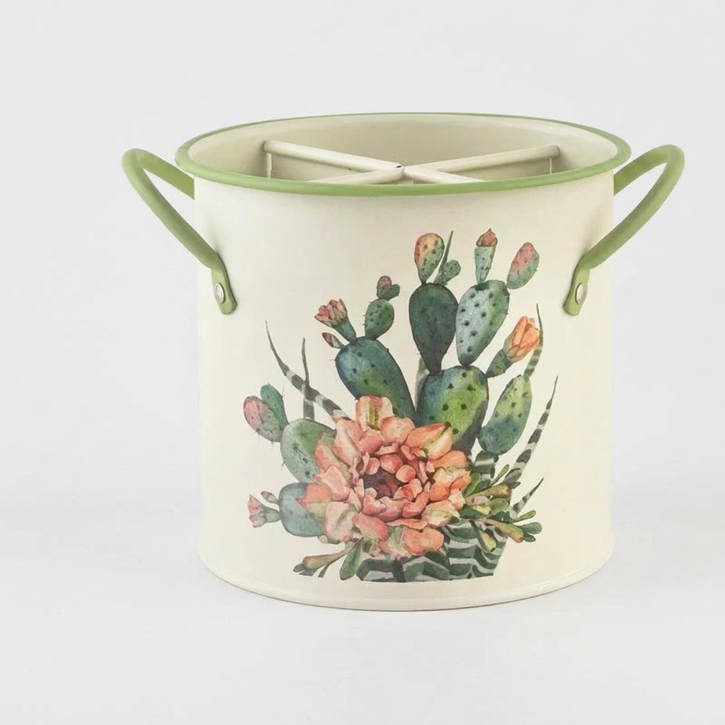 An off-white enamel bucket with green handles and a green rim, decorated with a watercolor illustration of a green prickly pear cactus and peach flowers. A white metal trivet is placed inside.
