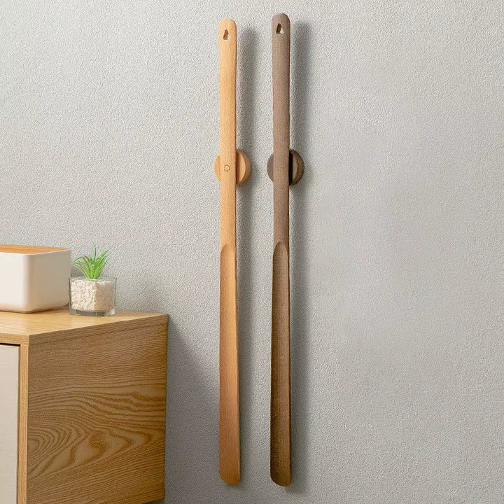 Two wooden brooms leaning against a light gray wall with a wooden shelf and plant in the corner.