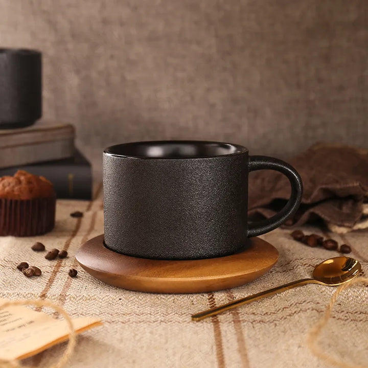 Black mug on a wooden coaster with coffee beans and a spoon on a textured surface.