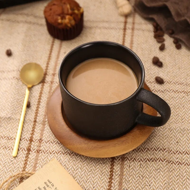 Black mug with coffee on a wooden coaster, surrounded by coffee beans and a muffin on a textured fabric background.