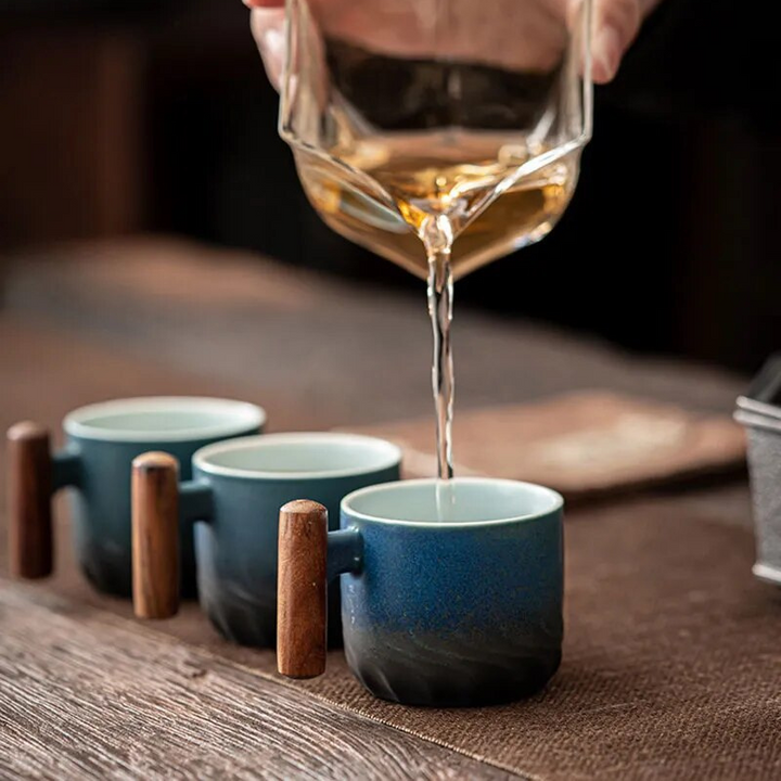 Tea being poured from a glass teapot into two blue ceramic cups on a wooden surface.