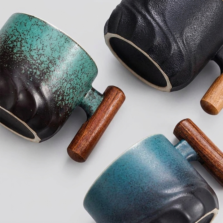 Three ceramic mugs with textured glazes in teal, black, and blue tones, each paired with a cylindrical wooden handle, arranged on a light gray background.