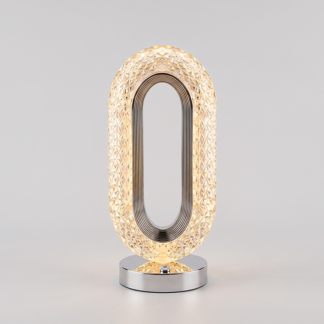 A modern oval table lamp with a textured, faceted clear glass shade that glows warm yellow, supported by a polished silver metal frame and a matching silver circular base, set against a neutral light gray background.