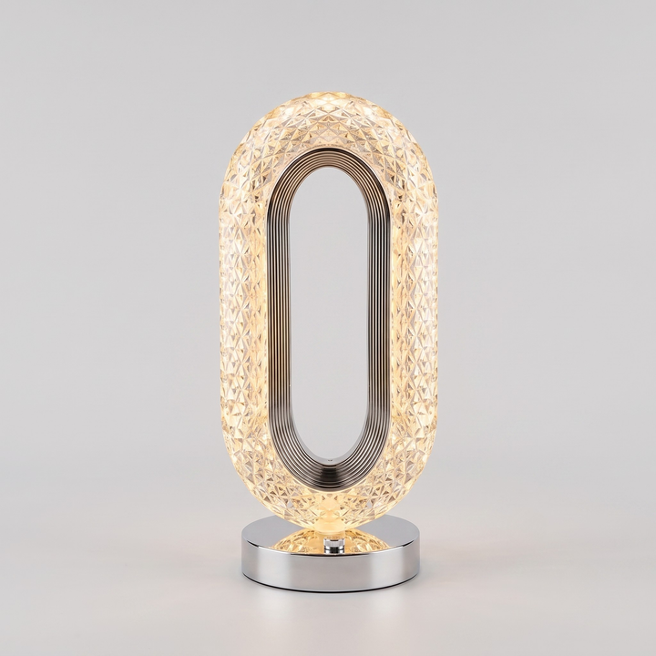 A modern oval table lamp with a textured, faceted clear glass shade that glows warm yellow, supported by a polished silver metal frame and a matching silver circular base, set against a neutral light gray background.