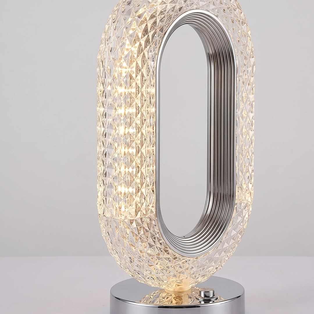 A modern oval table lamp with a clear faceted crystal-like textured body, a silver metallic inner frame, and a polished silver base, shown lit against a light gray background.