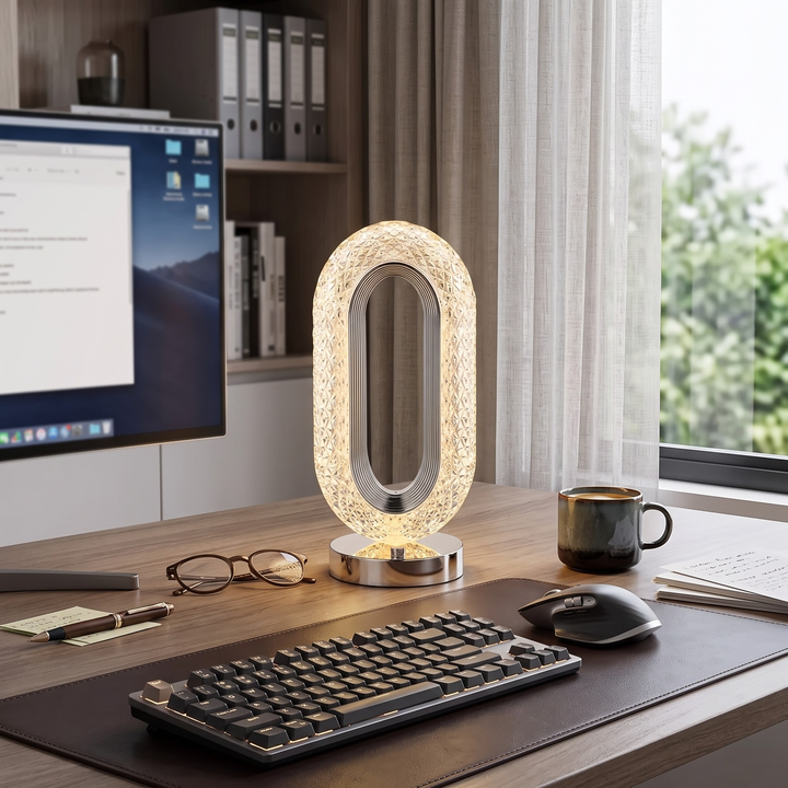 A modern oval LED desk lamp with a textured, crackled glass-like exterior and a polished silver base, illuminated on a wooden desk next to a keyboard, mouse, and computer monitor.