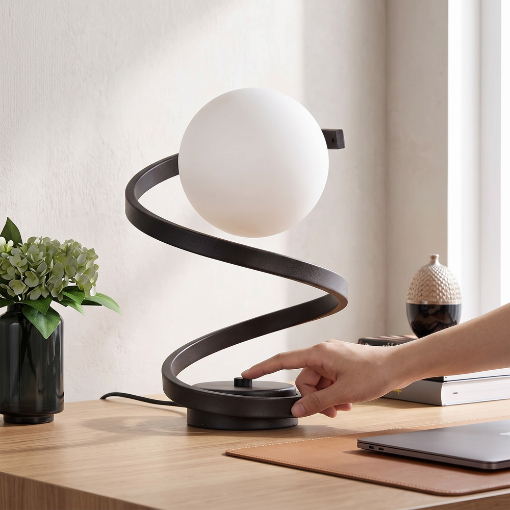 A modern spiral desk lamp with a matte black metal base and a white spherical shade, shown on a wooden desk with a hand turning its switch.