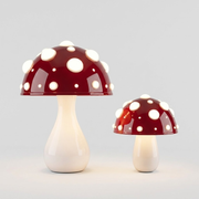 Two mushroom-shaped table lamps in different sizes, featuring glossy dark red caps with white spots and smooth off-white stems, set against a light gray background.