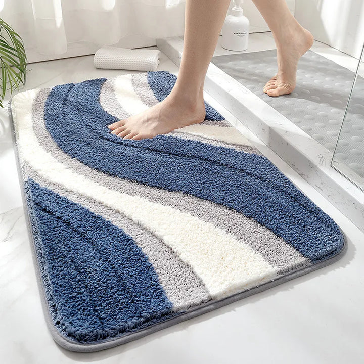 A rectangular bath mat with a plush, high-pile texture featuring wavy stripes of navy blue, medium blue, white, and light gray. A person's bare feet are stepping onto the mat in a bright, modern bathroom setting.