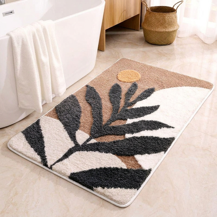 A rectangular bath mat with a modern botanical leaf pattern in black, beige, and white, styled on a beige tiled bathroom floor next to a white bathtub.