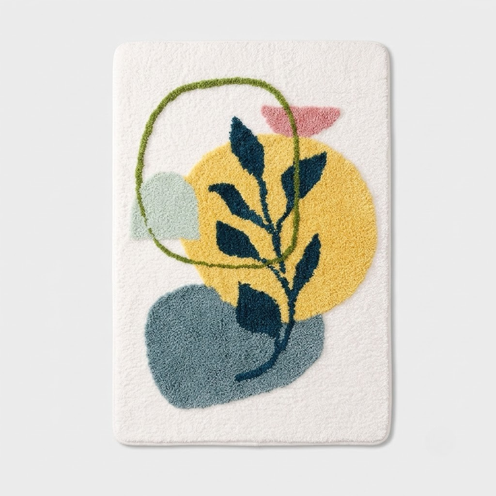 A rectangular white bath mat featuring a colorful abstract tufted design with a yellow circle, blue-green shape, pink shape, and dark green leafy branch, shown on a light gray background.