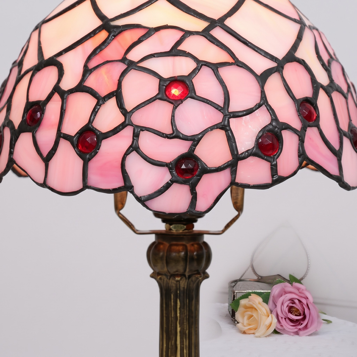 A Tiffany-style stained glass table lamp with a pink floral pattern, dark metal grid lines, and red crystal accents, shown lit against a light gray background with decorative roses at the base.