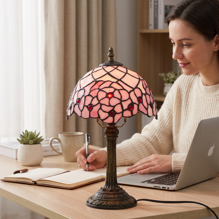 A Tiffany-style table lamp with a pink stained-glass shade and bronze base sits on a wooden desk in a home office setting, with a woman working on a laptop in the background.