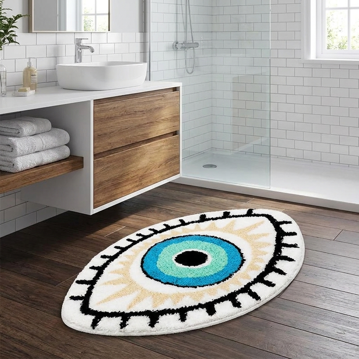 An oval bathroom rug with an eye design in blue, green, black, and beige, placed on a dark wood floor in a modern white-tiled bathroom.
