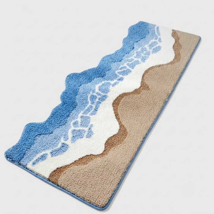 A rectangular bath mat with a beach and ocean wave design, featuring blue water, white foam, and beige sand on a light gray background.
