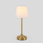 A modern table lamp with a tapered white fabric shade, a polished gold-finished metal stem, and a round gold base, shown illuminated against a neutral light gray background.