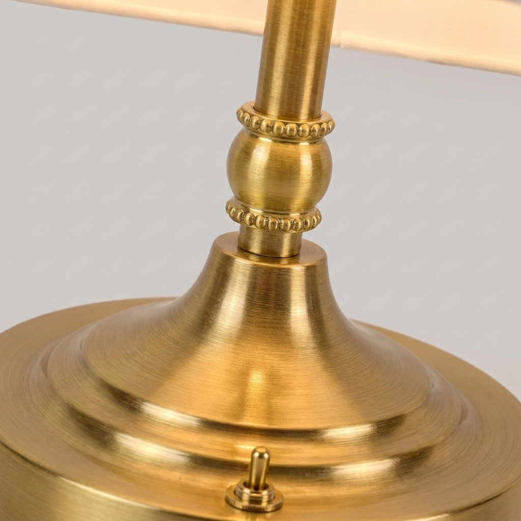 Close-up of a brass table lamp base and stem with a decorative beaded collar and a warm white fabric shade partially visible at the top, set against a neutral gray background.