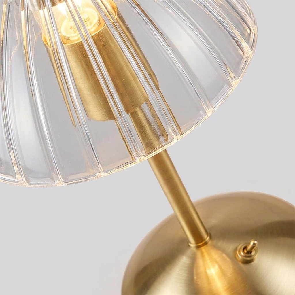 Close-up of a table lamp with a ribbed clear glass shade, a warm brass-toned stem, and a matching round base on a light gray background.