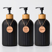 Three ribbed black pump bottles with light wood caps, arranged side by side on a white surface against a light gray background. Each bottle has a hanging wooden tag labeled Conditioner, Body Wash, and Shampoo.