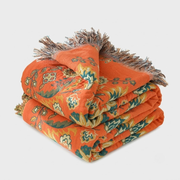 An orange throw blanket with a vintage floral pattern in green, yellow, and white, finished with brown fringed tassels, folded neatly on a light gray background.