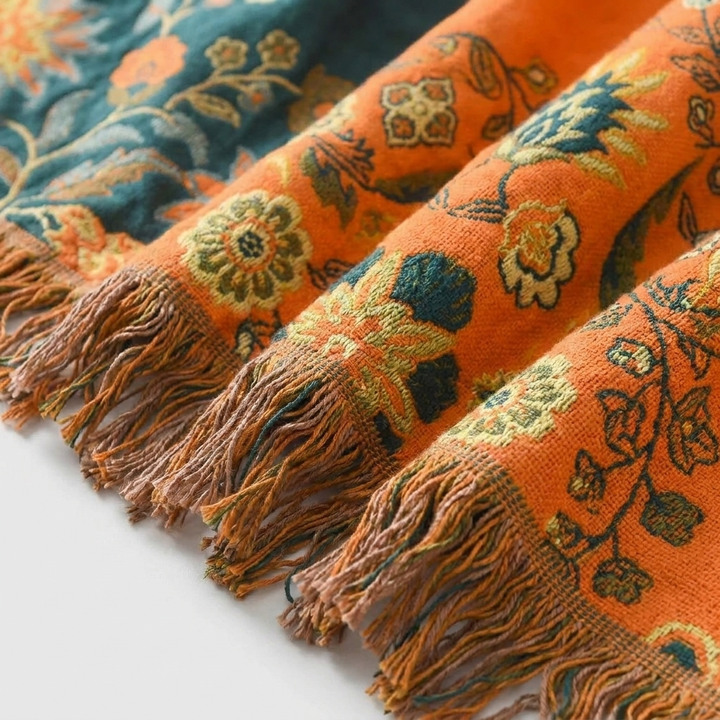 A close-up of a woven textile with fringe ends, featuring an orange base with floral and botanical patterns in teal, mustard yellow, and brown.