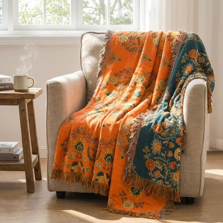 A vibrant orange and teal patterned throw blanket with fringe is draped over a beige armchair in a bright, sunlit room next to a small wooden side table with a mug.