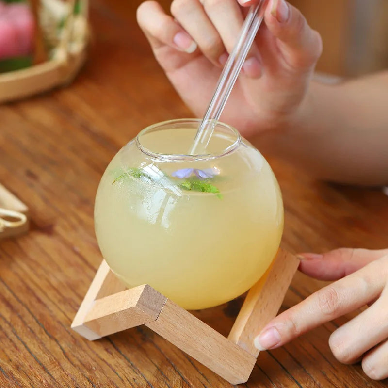 Glass ball with yellow liquid and a straw on a wooden stand, held by a person.