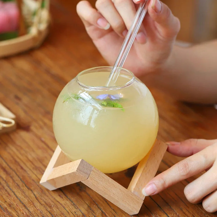 Glass ball with yellow liquid and a straw on a wooden stand, held by a person.