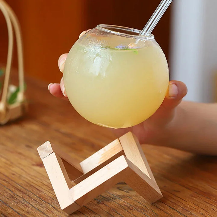 Hand holding a glass of yellow liquid with a straw on a wooden stand.