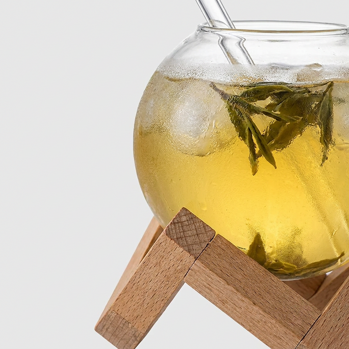 A clear glass teapot filled with pale yellow tea and visible green tea leaves, resting on a wooden stand against a light gray background.