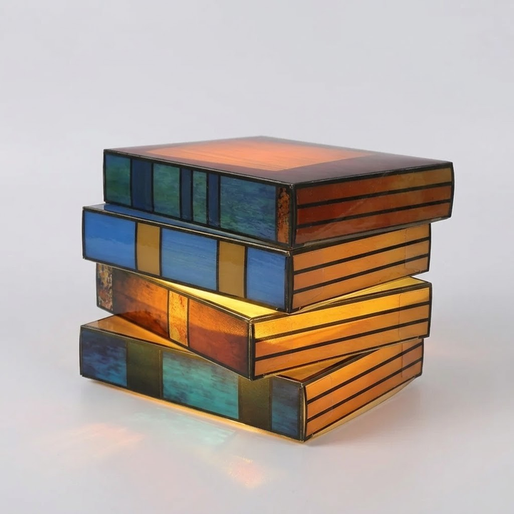 A decorative stack of four book-shaped lamps or boxes with stained-glass-style panels in amber, orange, blue, green, and gold tones, photographed on a clean light gray background.