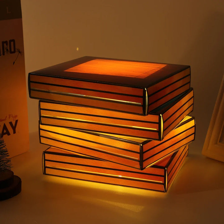A decorative lamp shaped like a stack of glowing books with warm amber and orange illumination, placed on a tabletop in a dim indoor setting.