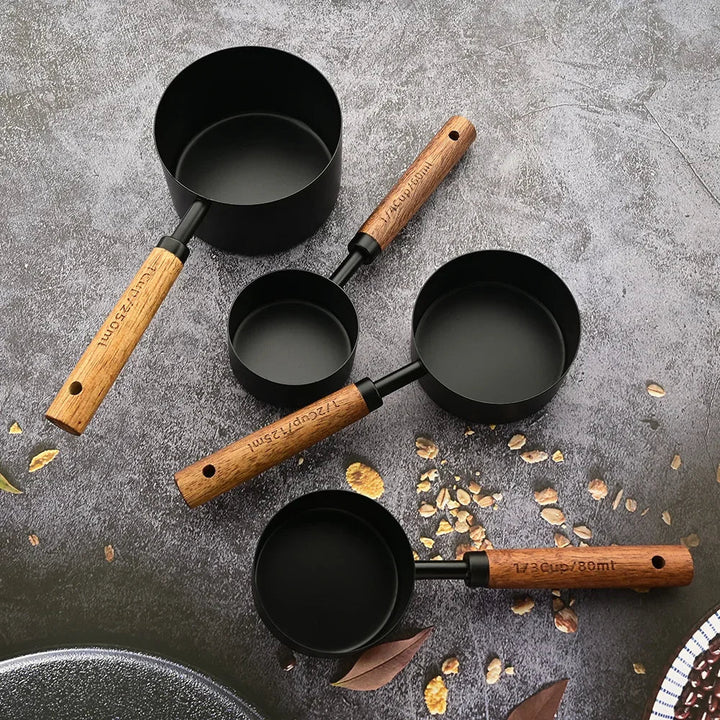 Set of black measuring cups with wooden handles on a gray surface with scattered leaves.