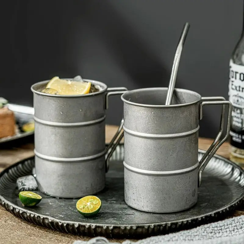 Two metal mugs with handles on a dark tray, containing lemon slices and a bottle of liquor in the background.