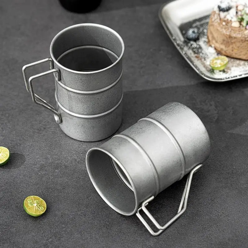 Two gray metal mugs with handles on a dark surface with limes and a plate of food.