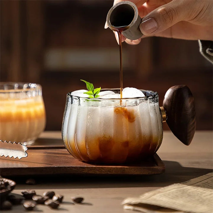 Person pouring a dark liquid into a glass of iced coffee with a wooden coaster and coffee beans on a wooden surface.