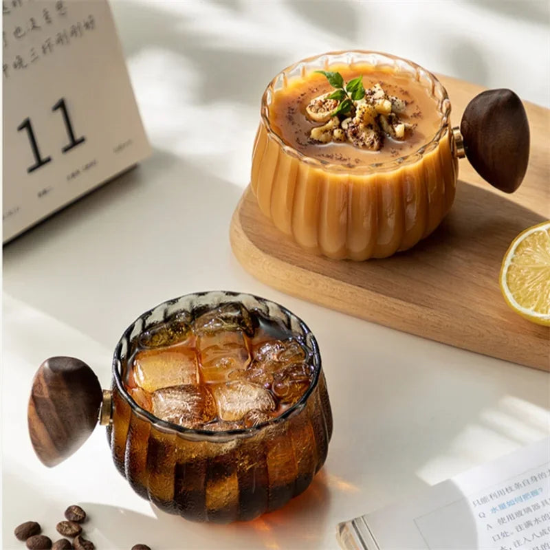 Two glass cups with wooden handles, one containing a dessert and the other iced coffee, on a light surface.