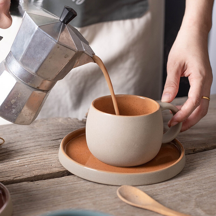 Person pouring coffee from a Moka pot into a ceramic cup on a wooden table.