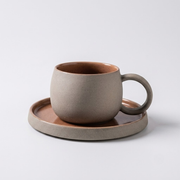 A matte ceramic coffee cup with a matching saucer, featuring a speckled taupe-gray exterior and a warm terracotta-brown interior, set against a clean light gray background.