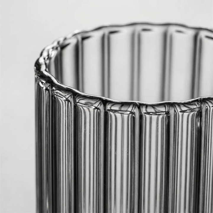 Close-up of a ribbed clear glass cup or vase with a scalloped rim, shown against a light gray background.