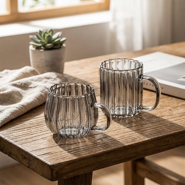 Two ribbed clear glass mugs with handles, placed on a rustic wooden table in a softly lit indoor setting with a small plant and folded cloth in the background.