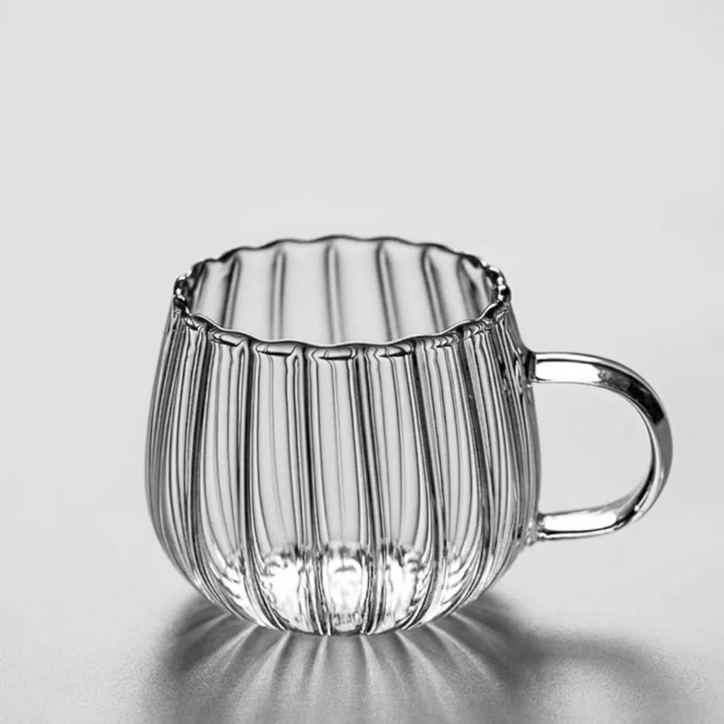 A clear ribbed glass mug with a scalloped rim and a curved handle, shown on a light gray reflective surface.