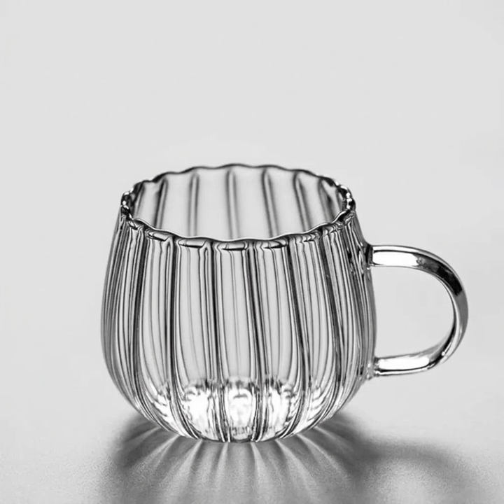 A clear ribbed glass mug with a scalloped rim and a curved handle, shown on a light gray reflective surface.